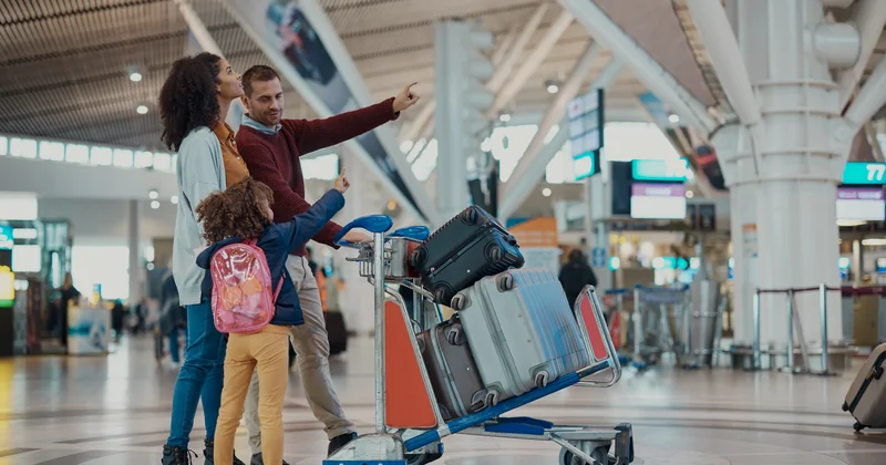 Familie cu bagaje într-un terminal de aeroport modern, privind panourile de îmbarcare - sejur charter TravelTheme